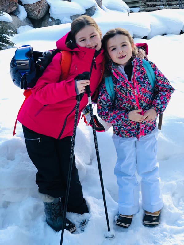 two young girls posing for photo on snow wearing ski outfits